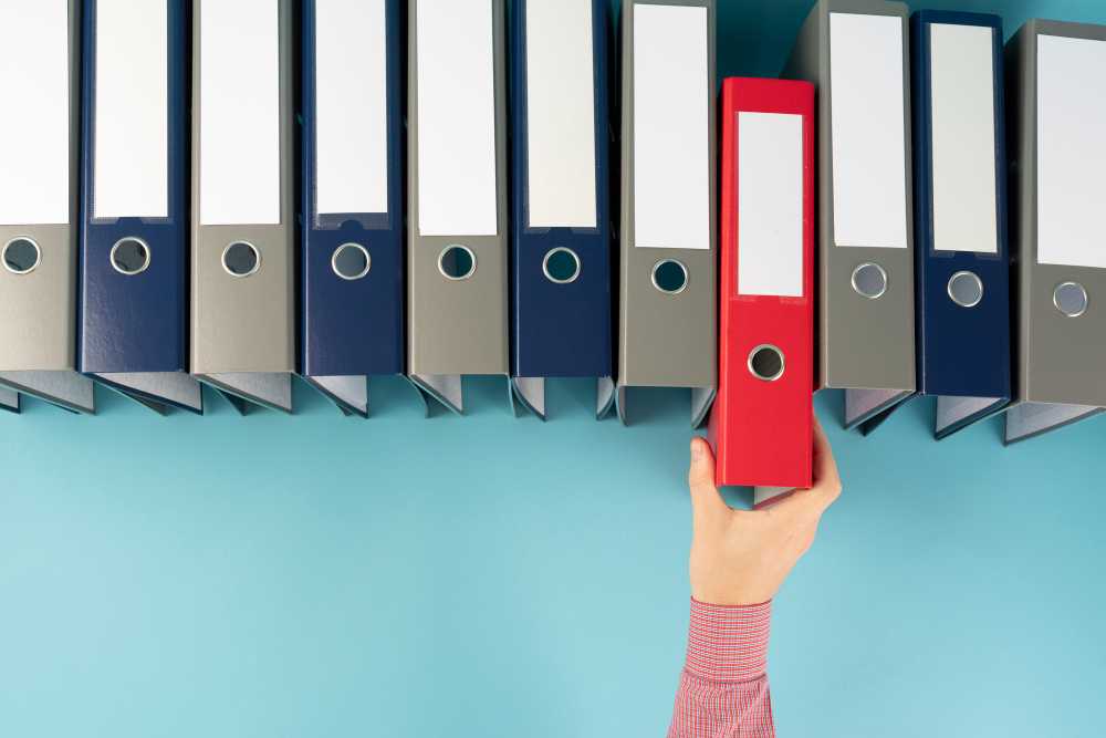 A row of office ring binders on a shelf, mostly blue and gray, with a hand pulling out a red binder, symbolizing records management and document organization.