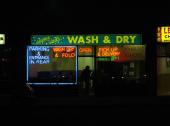 Night time photo of a wash & dry laundromat. A silhouette stands in the doorway with blue and red neon signs advertising available dry cleaning services