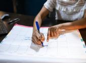 An event planner writes on a large desk calendar with a blue marker, using their other hand to indicate a specific date while organizing upcoming events.