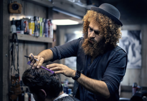 man with curly auburn hair and fedora cutting and making fine adjustment to a man's haircut