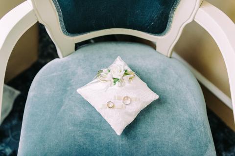 Two wedding rings sit on a small pillow on a blue chair.